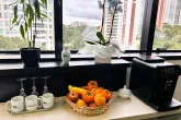 Imagem da mesa do café, na mesa tem uma cesta com frutas ao centro, canecas e taças no lado esquerdo da foto, cafeteira com um açucareiro do lado direito da foto. E vasos ao fundo na janela.  