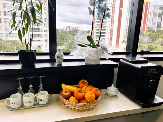 Imagem da mesa do café, na mesa tem uma cesta com frutas ao centro, canecas e taças no lado esquerdo da foto, cafeteira com um açucareiro do lado direito da foto. E vasos ao fundo na janela.  